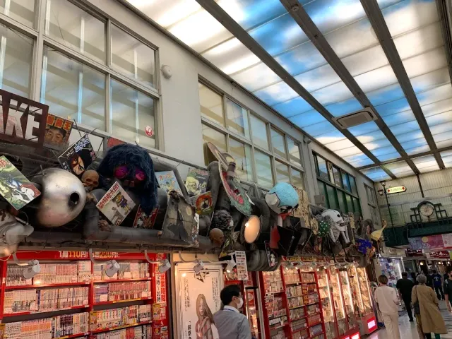 מעבר קניות מקורה עם חנות מנגה ופסלי מסכות מוזרות מעל חלונות הראווה