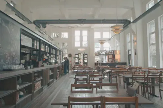 אולם גבוה ומואר עם דלפק ארוך, שולחנות וכיסאות עץ וחלונות גבוהים